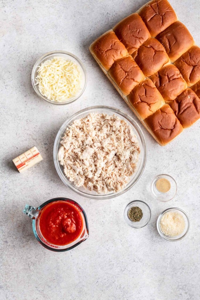 shredded chicken in a bowl surrounded by other ingredients like slider buns, marinara sauce and cheese