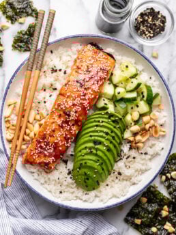 Salmon bowl with rice, cucumber, avocado, and seaweed on a white plate, garnished with sesame seeds.