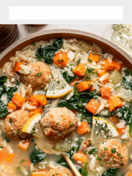 A bowl of lemony chicken meatball soup with spinach, carrots, celery, orzo pasta, lemon slices, and fresh herbs, shown from above with a spoon in the bowl.