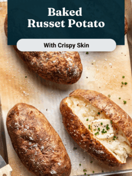 Four baked russet potatoes with crispy, salted skins rest on a parchment-lined baking sheet. One potato is cut open, revealing a fluffy interior topped with chopped chives. A fork and knife are nearby.