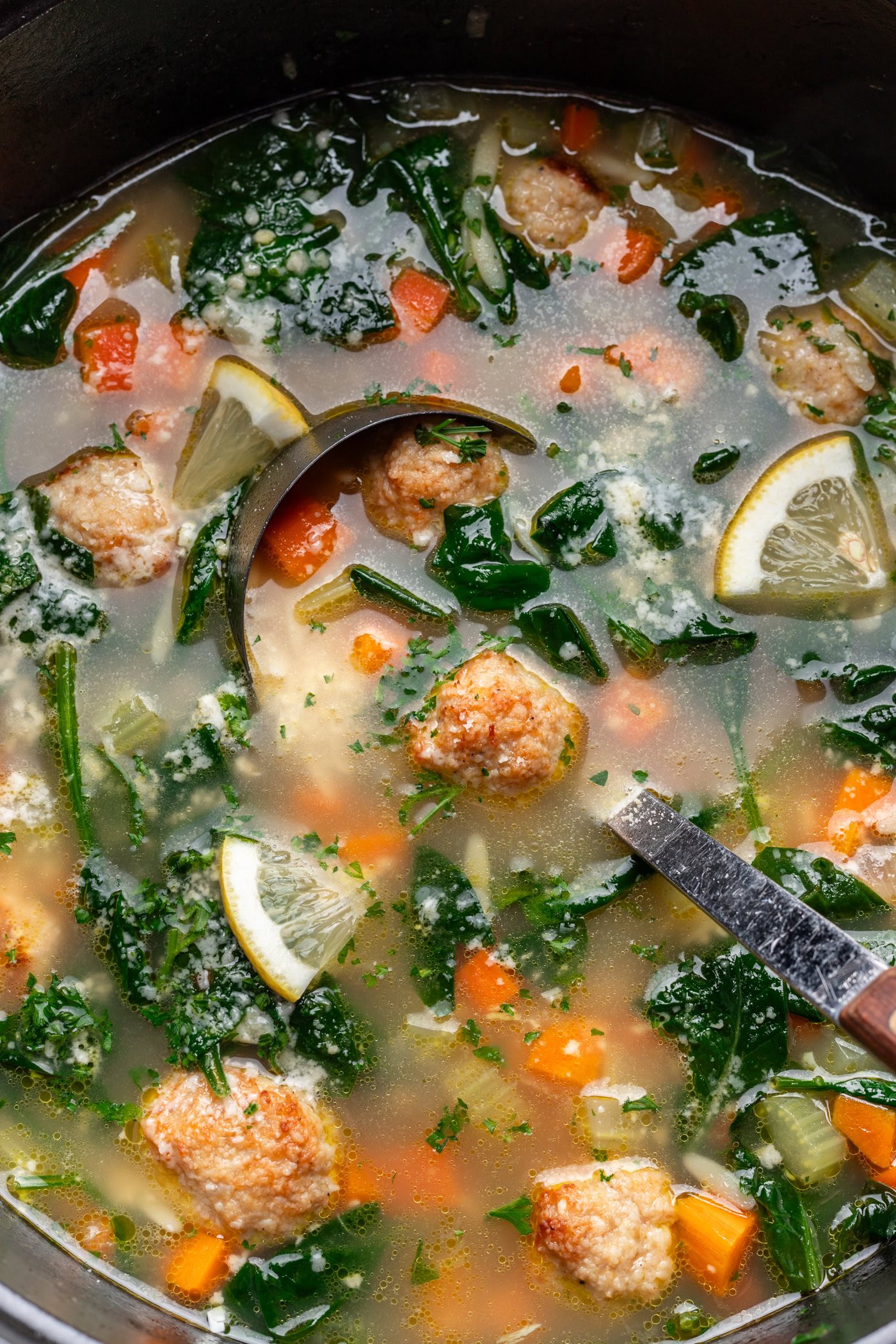 a ladle dipping into a big pot of soup with veggies, meatballs and greens
