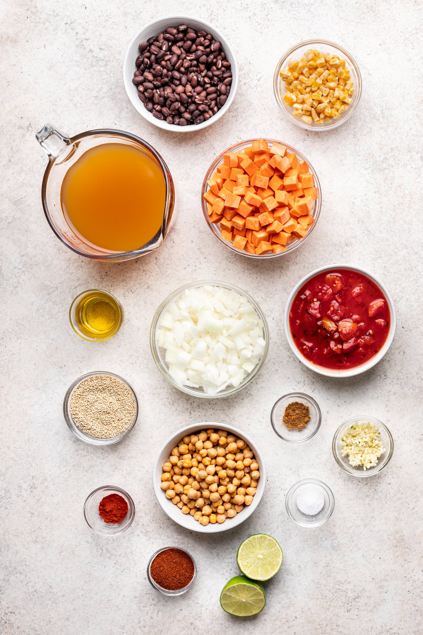 Overhead view of various ingredients in bowls on a light surface, including black beans, corn, diced sweet potatoes, vegetable broth, olive oil, onion, canned tomatoes, quinoa, chickpeas, spices, garlic, and lime halves.