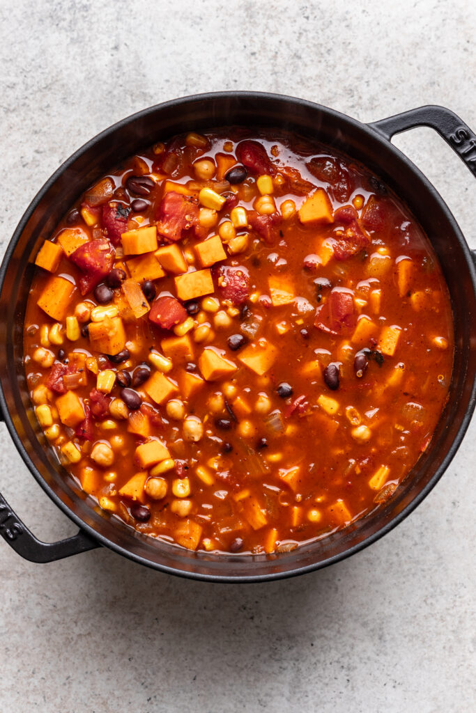 A pot of hearty vegetable chili with black beans, chickpeas, corn, diced tomatoes, and chunks of sweet potato in a rich, red broth, viewed from above.