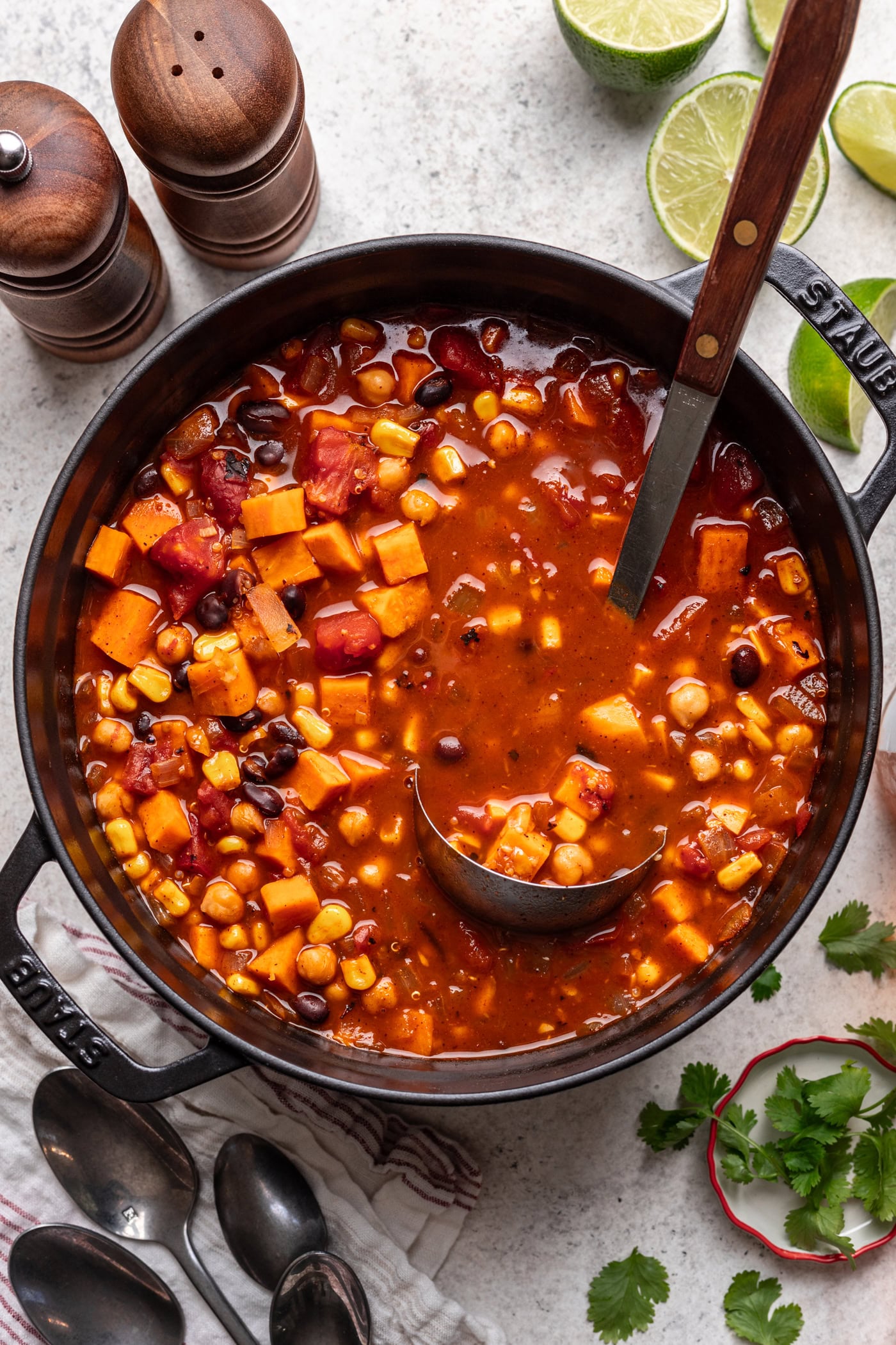 A pot of hearty vegetable chili with beans, corn, tomatoes, and sweet potatoes in a rich broth. A ladle rests inside the pot, surrounded by lime halves, fresh cilantro, spoons, and salt and pepper shakers.