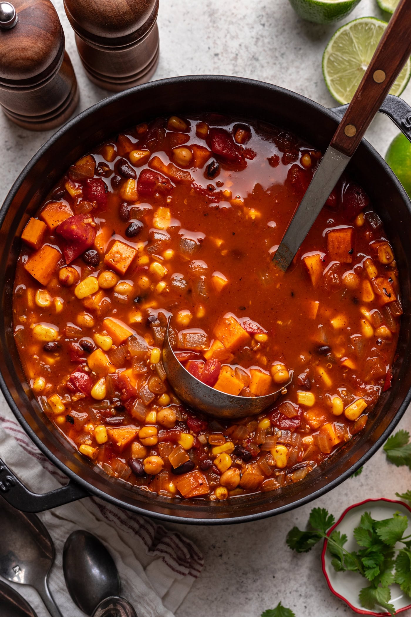 A pot of vibrant chili featuring corn, black beans, diced tomatoes, and sweet potatoes, with a ladle inside. Fresh limes, cilantro, and pepper mills are nearby on a light countertop.