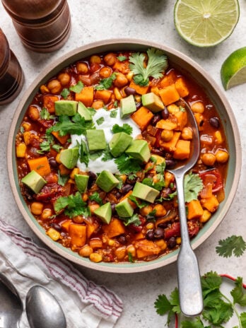 A bowl of vegetarian chili with sweet potatoes, black beans, corn, and chickpeas, topped with diced avocado, sour cream, and fresh cilantro, with a spoon inside and lime wedges and napkin nearby.