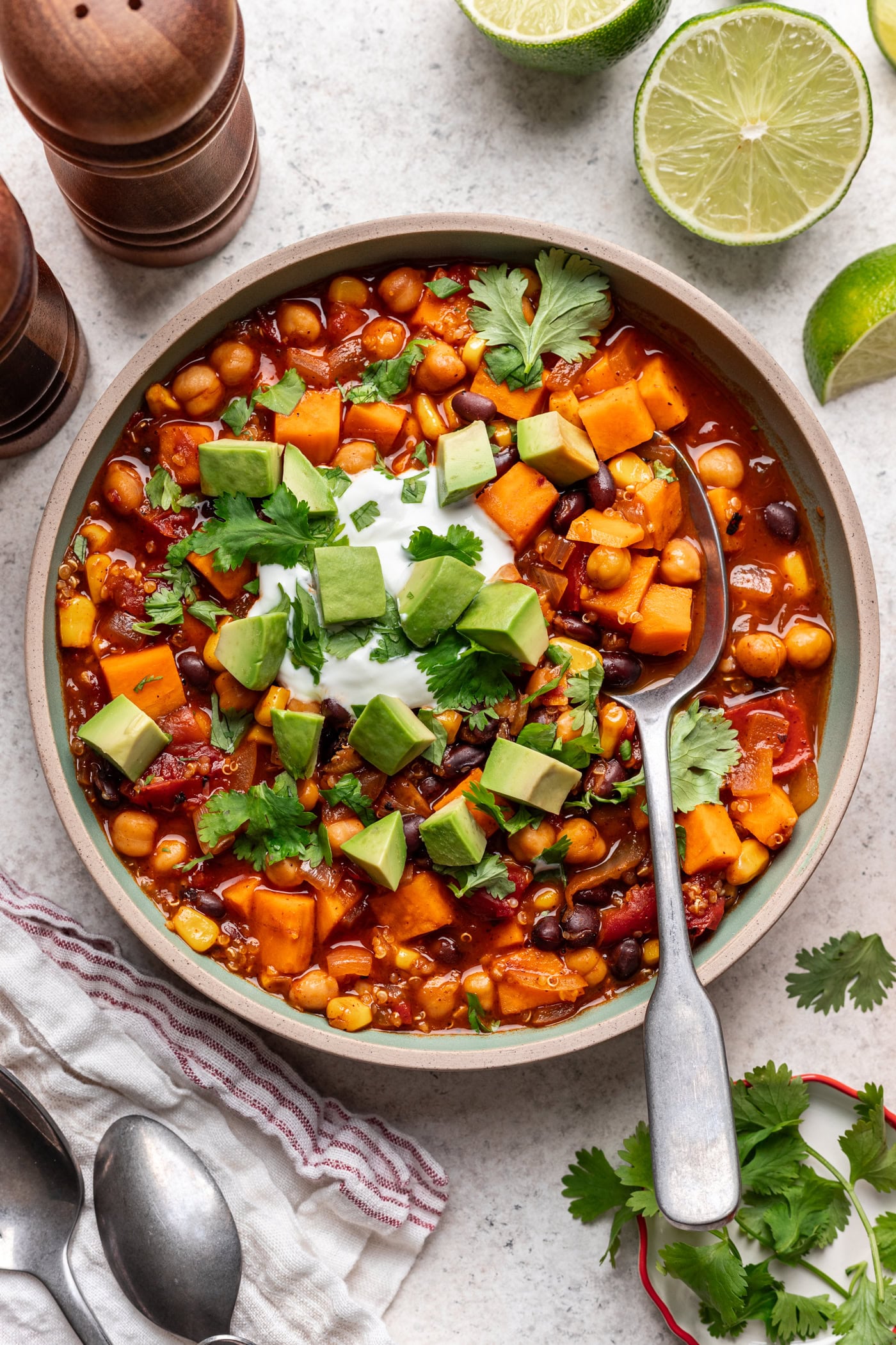 A bowl of vegetarian chili with sweet potatoes, black beans, corn, and chickpeas, topped with diced avocado, sour cream, and fresh cilantro, with a spoon inside and lime wedges and napkin nearby.