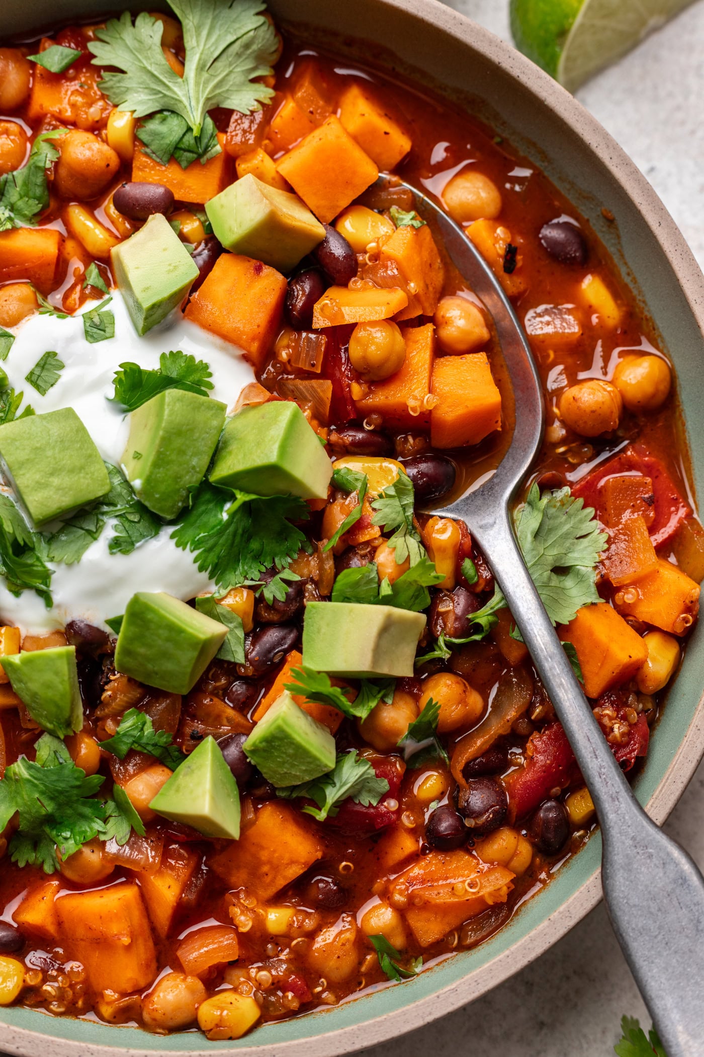 A close-up of a vibrant vegetable chili topped with diced avocado, fresh cilantro, and a dollop of sour cream. Ingredients like sweet potato, chickpeas, black beans, and corn are visible, with a spoon resting in the bowl.