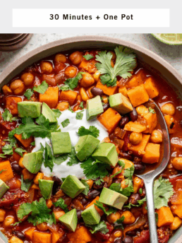 A bowl of sweet potato black bean chili with quinoa, topped with avocado cubes, fresh cilantro, and a dollop of sour cream. A spoon rests in the bowl and lime wedges are on the side, highlighting this hearty sweet potato black bean chili.
