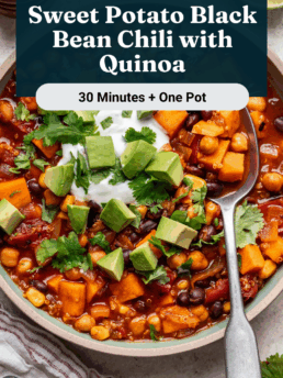 A bowl of hearty sweet potato black bean chili with quinoa, topped with diced avocado, cilantro, and sour cream, sits beside lime slices and a wooden spoon. Text overlay reads: “Sweet Potato Black Bean Chili with Quinoa.”.