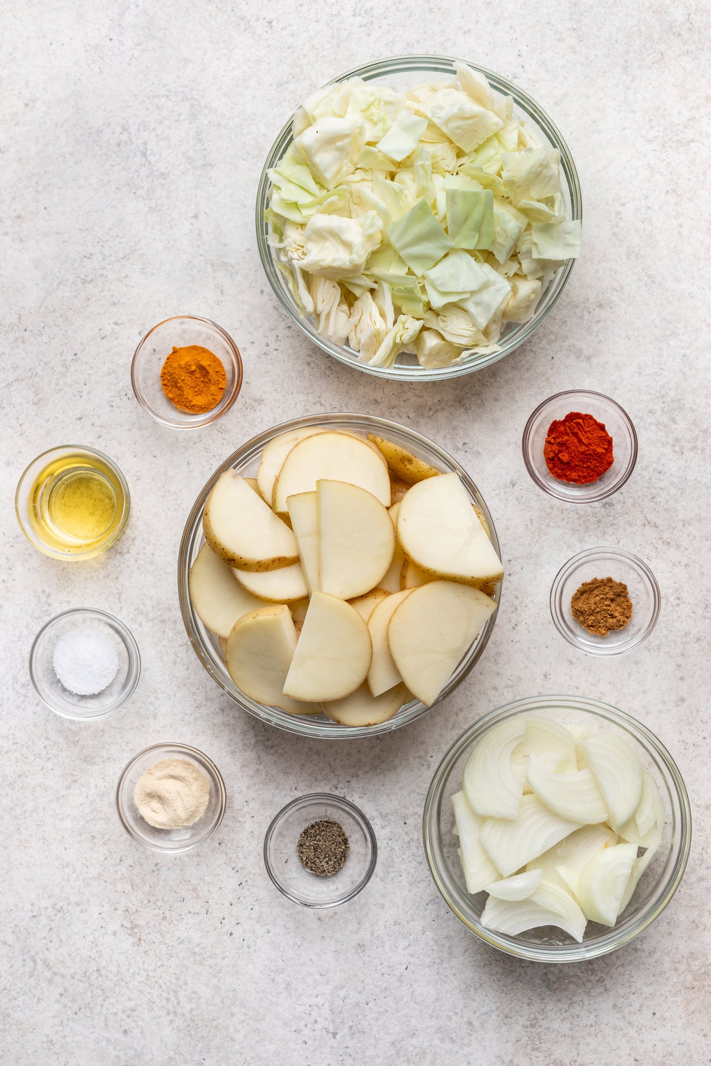 Three glass bowls filled with chopped cabbage, sliced potatoes and sliced onions, surrounded by small bowls of spices, salt, pepper and oil on a light countertop.