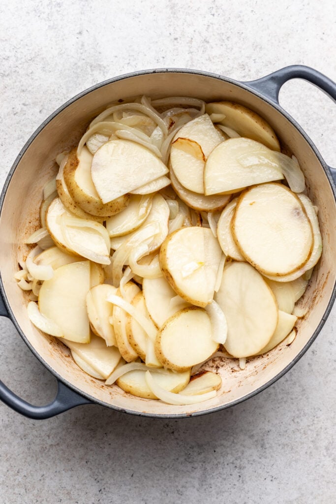 A round, gray pot filled with sliced potatoes and onions, lightly browned, sits on a light-colored countertop.