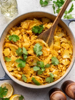 A pot of cooked cabbage and potato garnished with fresh cilantro, with a wooden spoon inside. Fresh lemon slices, cilantro, salt and pepper shakers, and a glass bottle are nearby on a light surface.