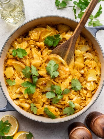 A pot of cooked cabbage and potato garnished with fresh cilantro, with a wooden spoon inside. Fresh lemon slices, cilantro, salt and pepper shakers, and a glass bottle are nearby on a light surface.