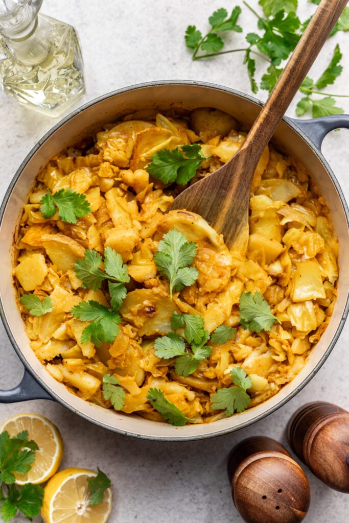 A pot of cooked cabbage and potato garnished with fresh cilantro, with a wooden spoon inside. Fresh lemon slices, cilantro, salt and pepper shakers, and a glass bottle are nearby on a light surface.