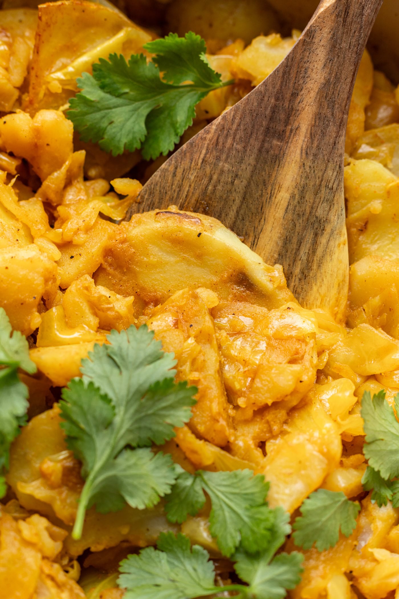 Close-up of a wooden spoon stirring a dish of cooked potatoes with a yellow seasoning, garnished with fresh cilantro leaves.