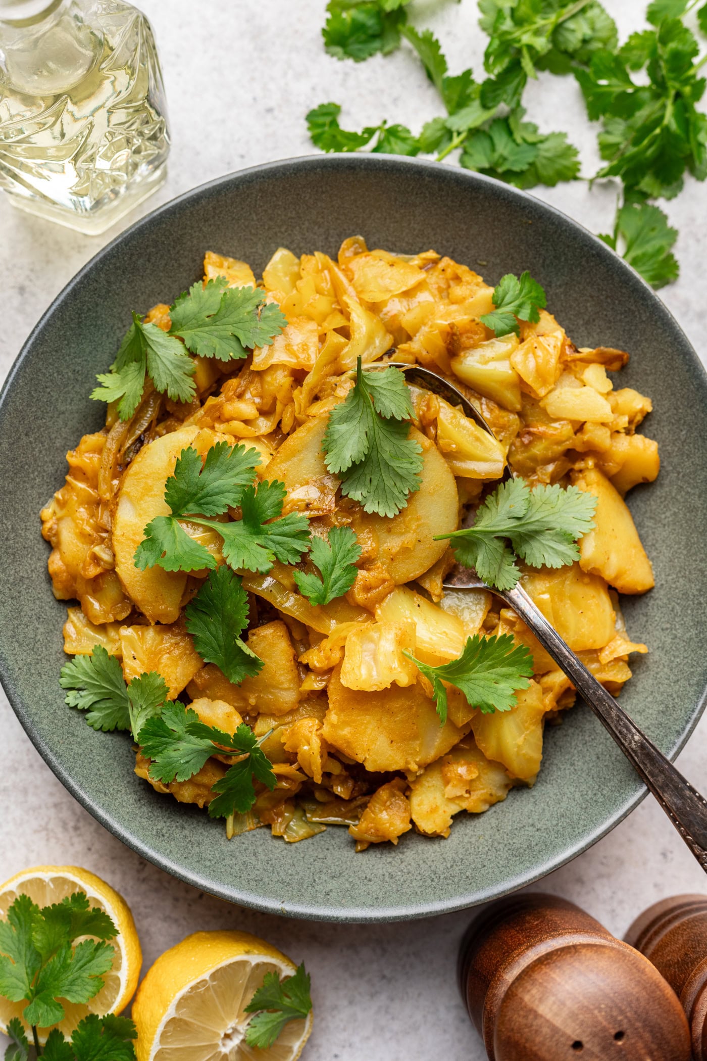 A bowl of cooked spiced potatoes garnished with fresh cilantro, with a metal spoon resting inside. Surrounding the bowl are lemon halves, more cilantro, a glass bottle, and wooden salt and pepper shakers.