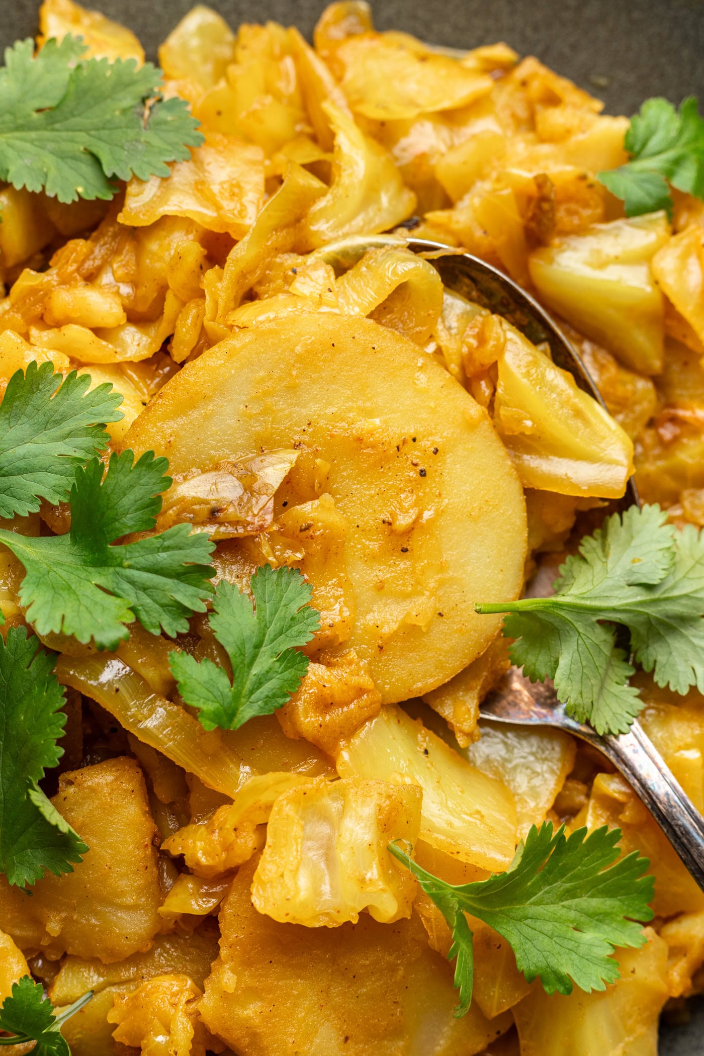Close-up of a dish featuring sliced potatoes and cabbage cooked with spices, garnished with fresh cilantro leaves. A spoon is partially visible among the ingredients.
