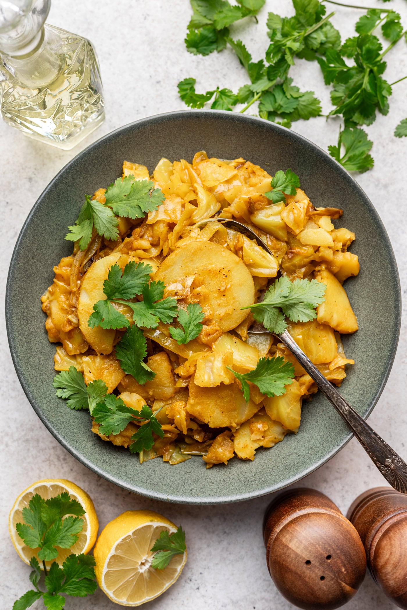 A bowl of spiced potato and cabbage garnished with fresh cilantro, next to lemon halves, fresh cilantro sprigs, a bottle of oil, and salt and pepper shakers on a light surface.