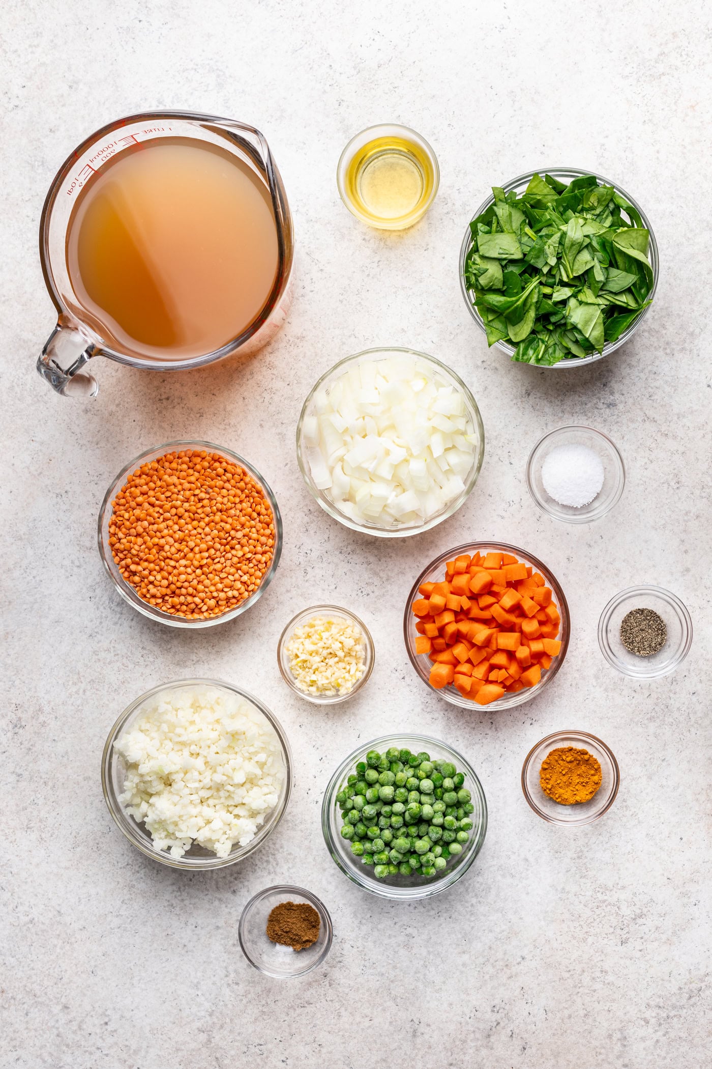 Overhead view of bowls containing vegetable broth, olive oil, chopped spinach, chopped onion, red lentils, diced carrot, salt, pepper, chopped garlic, chopped cauliflower, green peas, turmeric and cumin.