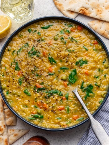 A bowl of yellow lentil soup with carrots, peas, and spinach, topped with cracked black pepper. Slices of flatbread, a lemon wedge, and a glass of water are nearby on a light surface. A spoon rests in the bowl.