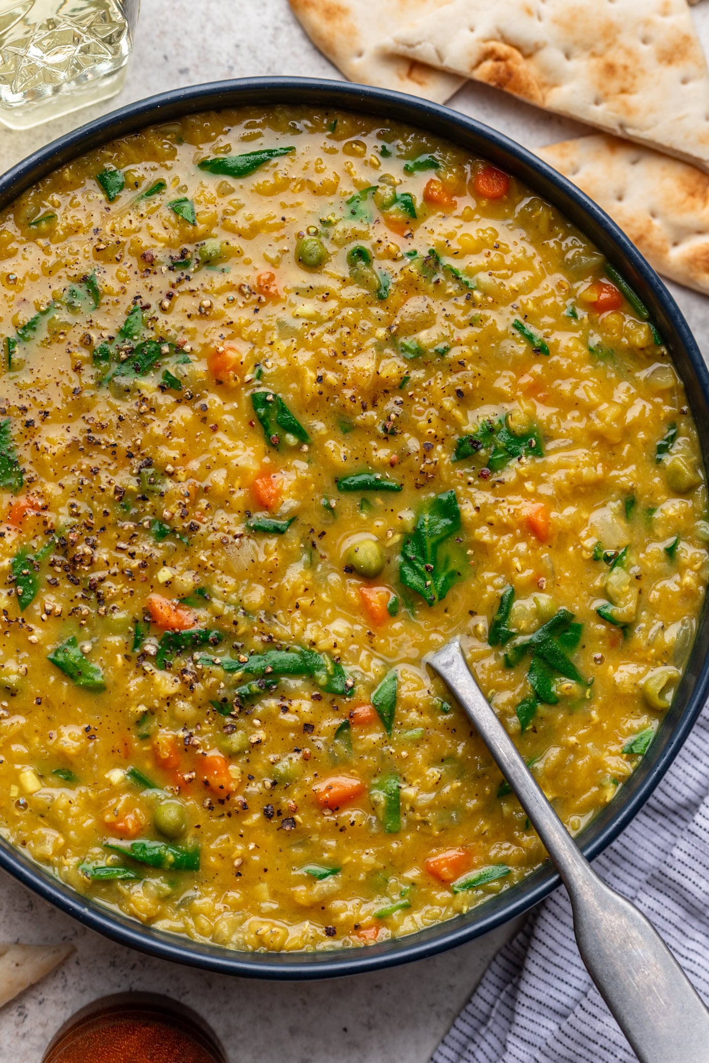 A bowl of thick yellow lentil soup with visible diced carrots, green peas, spinach, and black pepper on top, served with a spoon and pieces of flatbread on the side.