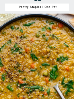 A bowl of thick red lentil soup with visible carrots, peas, greens, and black pepper on top. A spoon is in the bowl, and a glass of water is nearby. The text describes the soup as a one-pot meal with pantry staples.