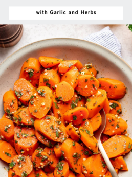 A bowl filled with steamed carrot slices garnished with chopped herbs and garlic. A silver spoon rests in the bowl. The text above reads Steamed Carrot Coins with Garlic and Herbs.