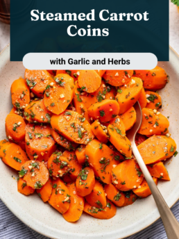 A white plate filled with sliced steamed carrots garnished with chopped herbs and garlic, with a silver spoon, a lemon half, and a pepper mill nearby on a light surface. Text on the image reads: Steamed Carrot Coins with Garlic and Herbs.