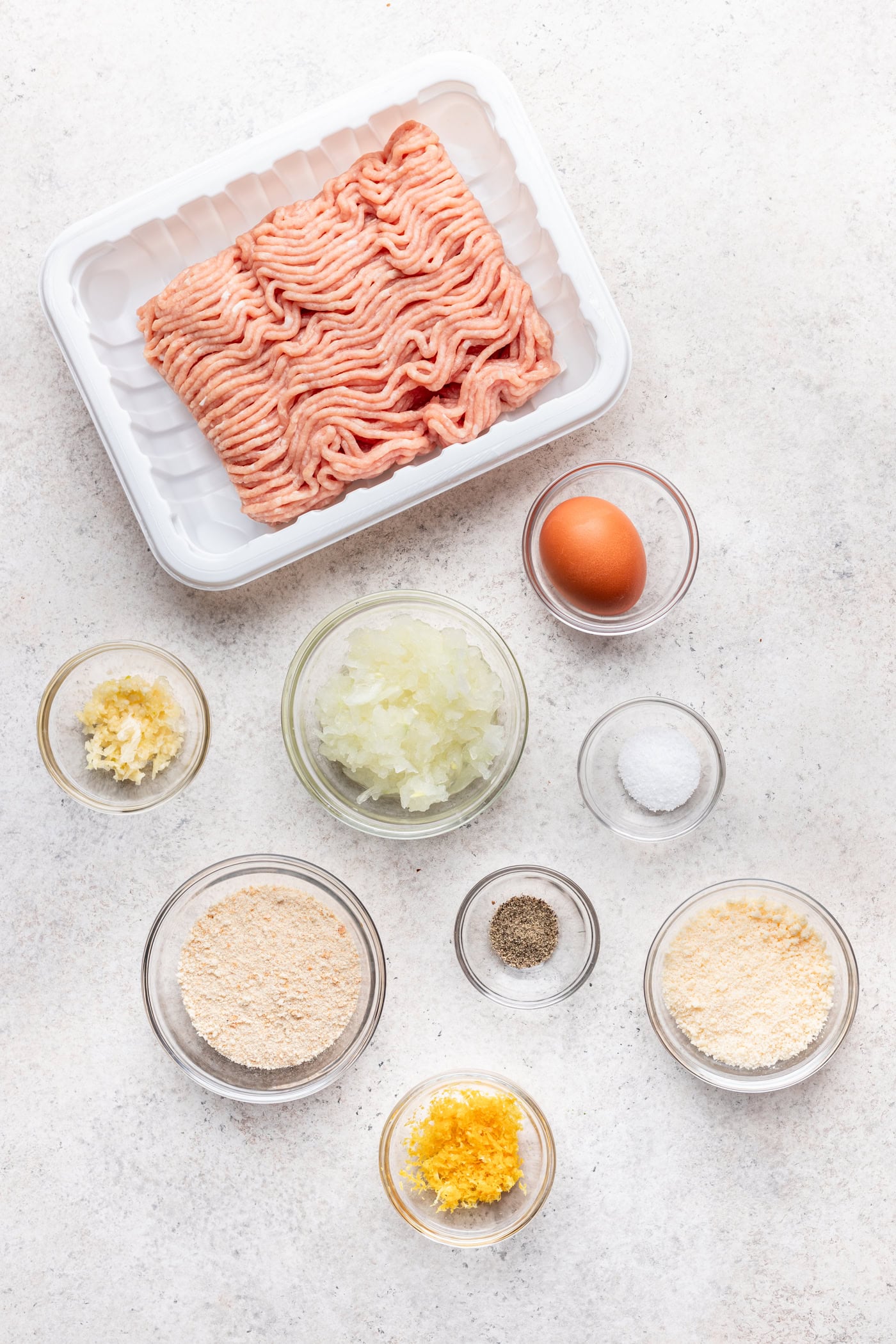 Top-down view of raw ground meat in a white tray, with small bowls containing an egg, minced garlic, chopped onion, salt, pepper, breadcrumbs, grated cheese, and lemon zest on a light countertop.