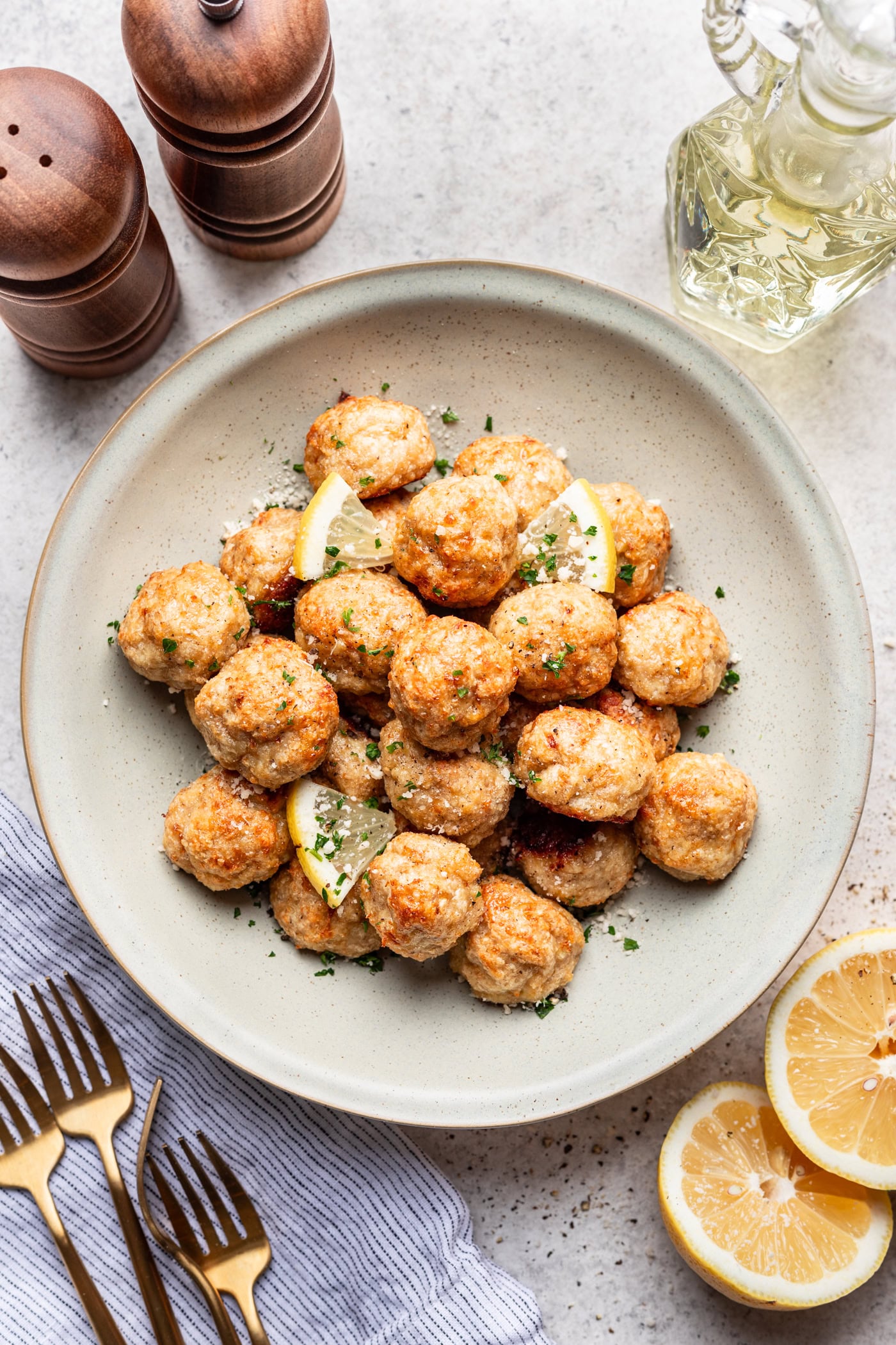 A bowl of seasoned meatballs garnished with chopped herbs and lemon wedges, surrounded by lemon halves, gold forks, a napkin, pepper mills, and a glass bottle of oil on a light surface.