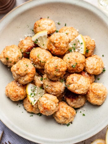 A plate of baked meatballs garnished with chopped herbs, grated cheese, and lemon wedges. The dish is served on a light-colored plate on a white surface.