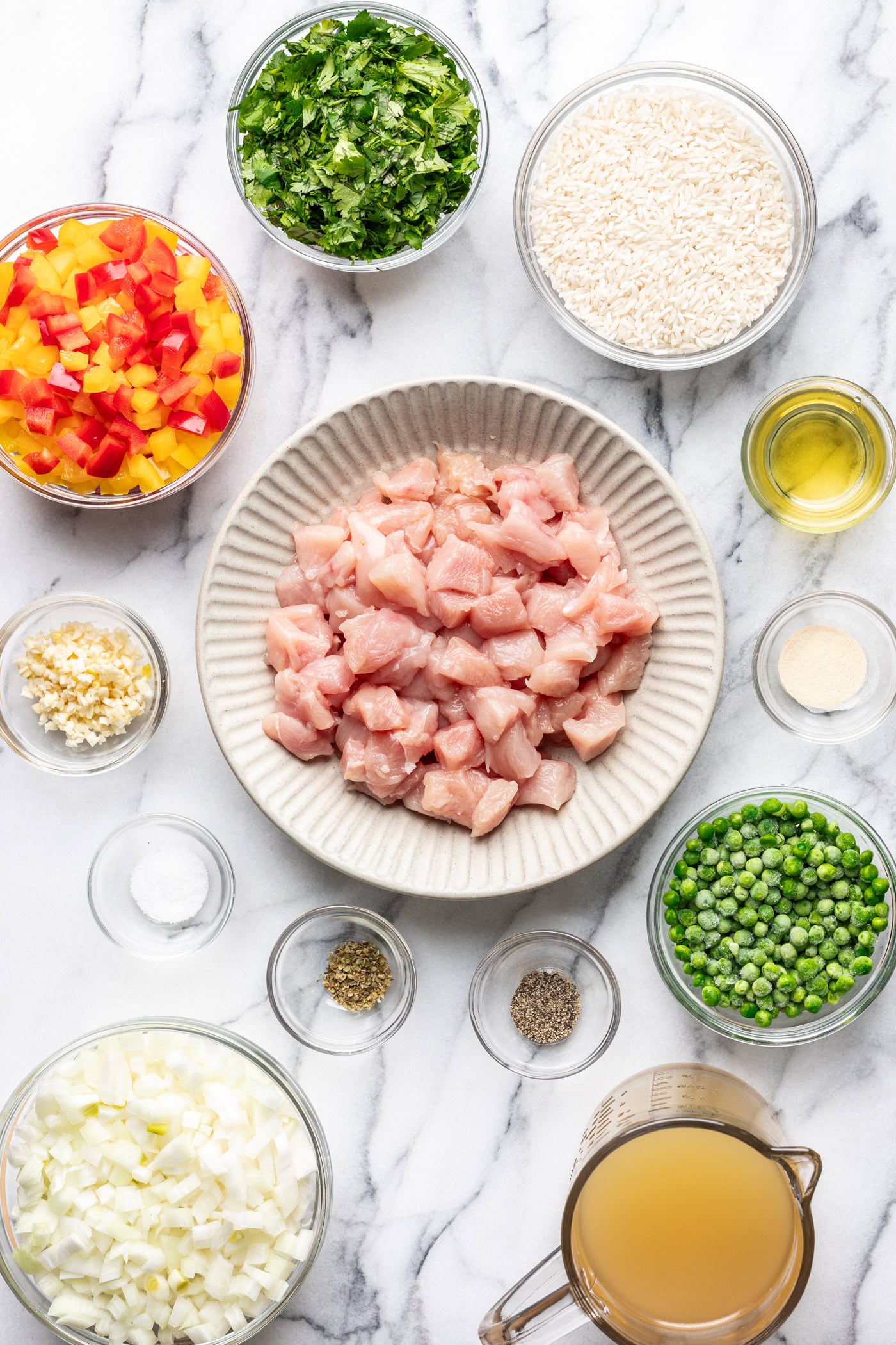 Overhead view of diced raw chicken on a plate surrounded by bowls of chopped bell peppers, rice, parsley, peas, diced onion, minced garlic, broth, oil, and seasonings on a marble surface.