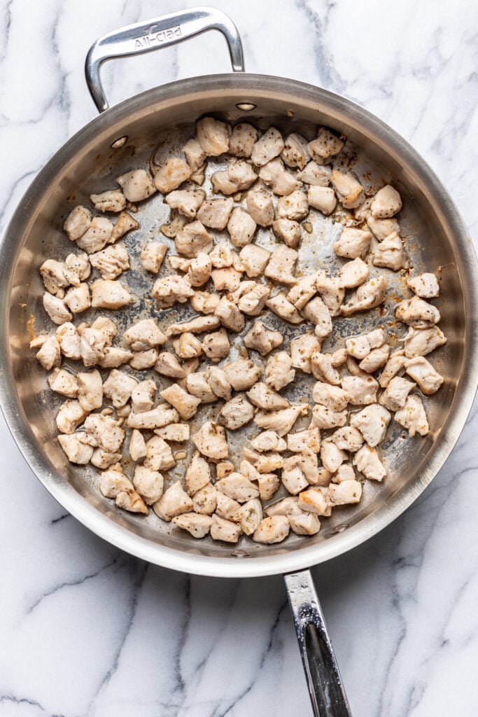 A stainless steel skillet filled with cubed, cooked chicken pieces seasoned with pepper, resting on a white marble countertop.