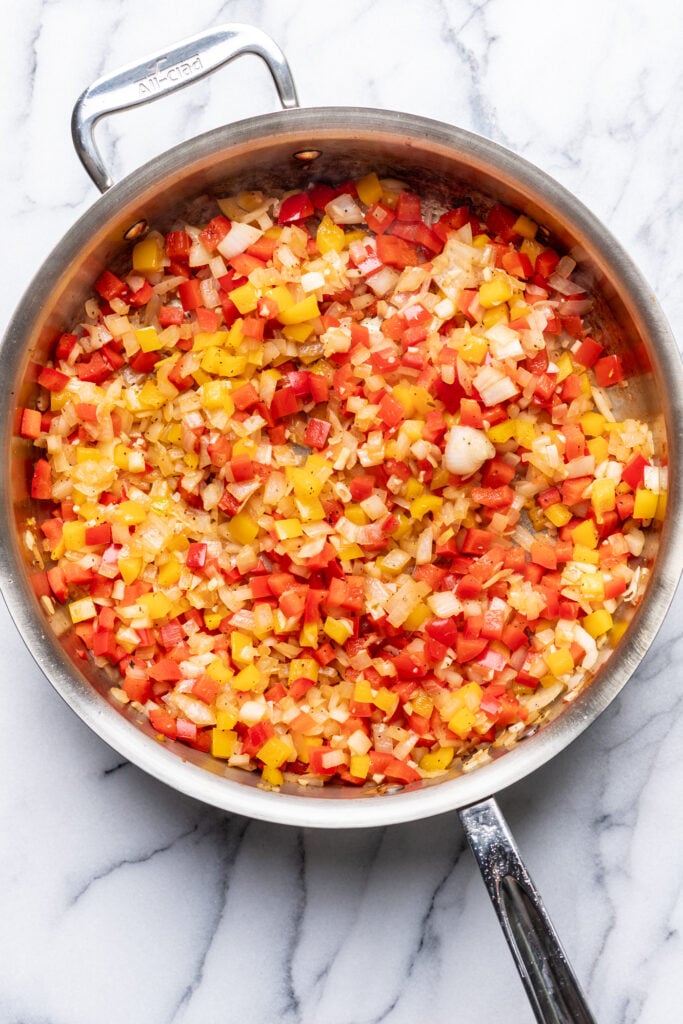 A stainless steel pan on a marble surface filled with sautéed diced red and yellow bell peppers, onions, and garlic.