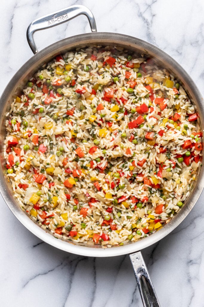 A stainless steel pan filled with cooked rice mixed with diced red, yellow, and green bell peppers, and green peas, sitting on a white marble surface.