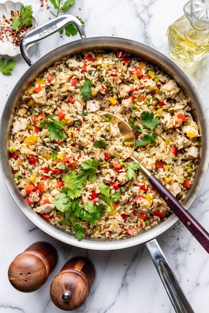 A skillet filled with cooked rice, diced chicken, and colorful bell peppers, garnished with fresh cilantro. A serving spoon rests in the pan. Salt and pepper shakers and a bottle of oil are nearby on a marble countertop.