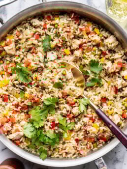 A skillet filled with cooked rice mixed with diced chicken, red and yellow bell peppers, peas, and garnished with fresh cilantro. A serving spoon rests in the skillet on a marble surface.