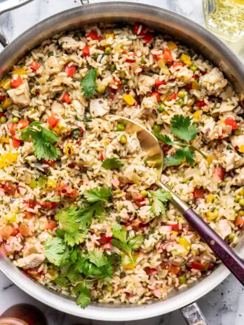 A skillet filled with cooked rice mixed with diced chicken, red and yellow bell peppers, peas, and garnished with fresh cilantro. A serving spoon rests in the skillet on a marble surface.
