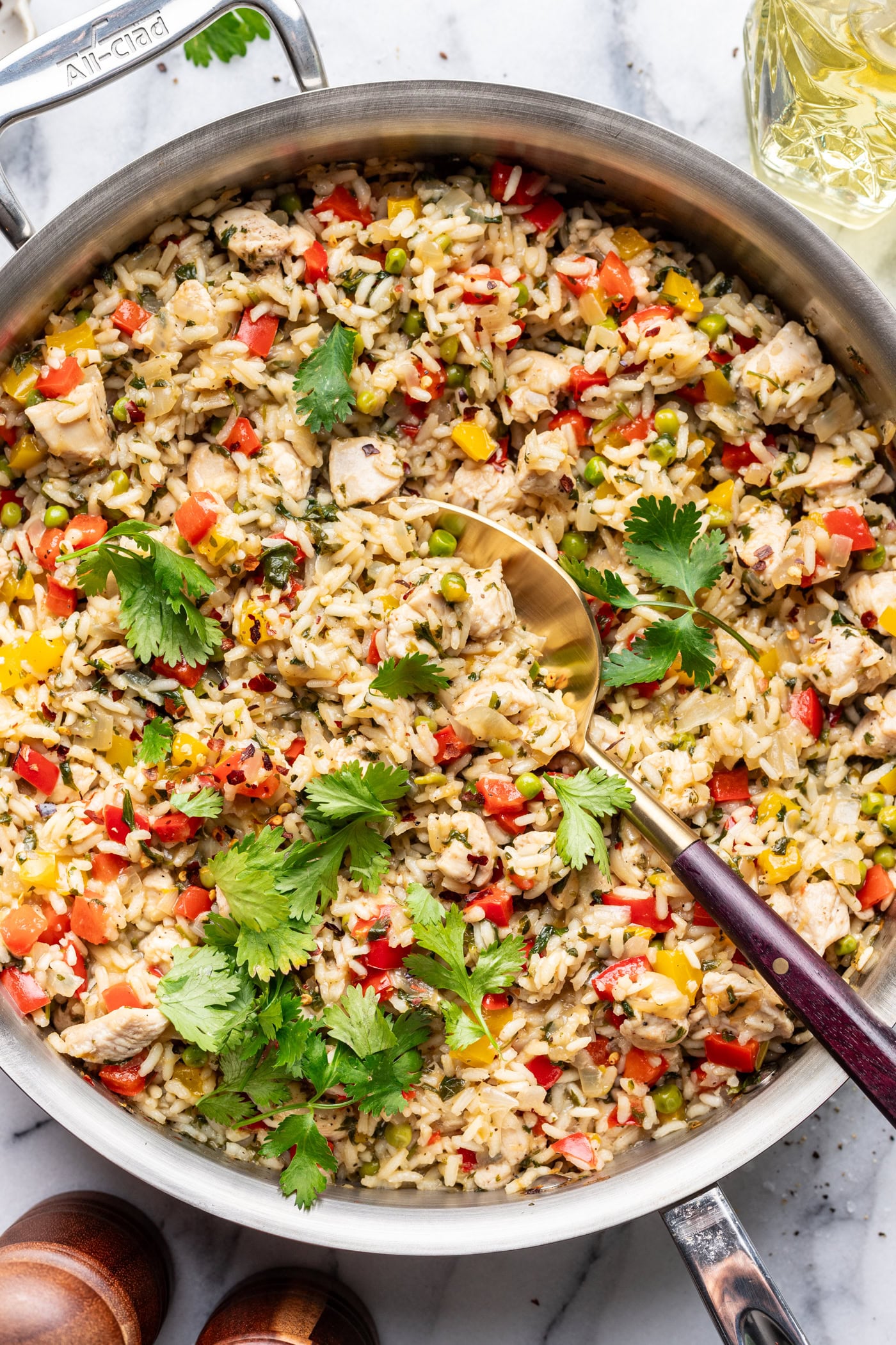 A skillet filled with cooked rice mixed with diced chicken, red and yellow bell peppers, peas, and garnished with fresh cilantro. A serving spoon rests in the skillet on a marble surface.