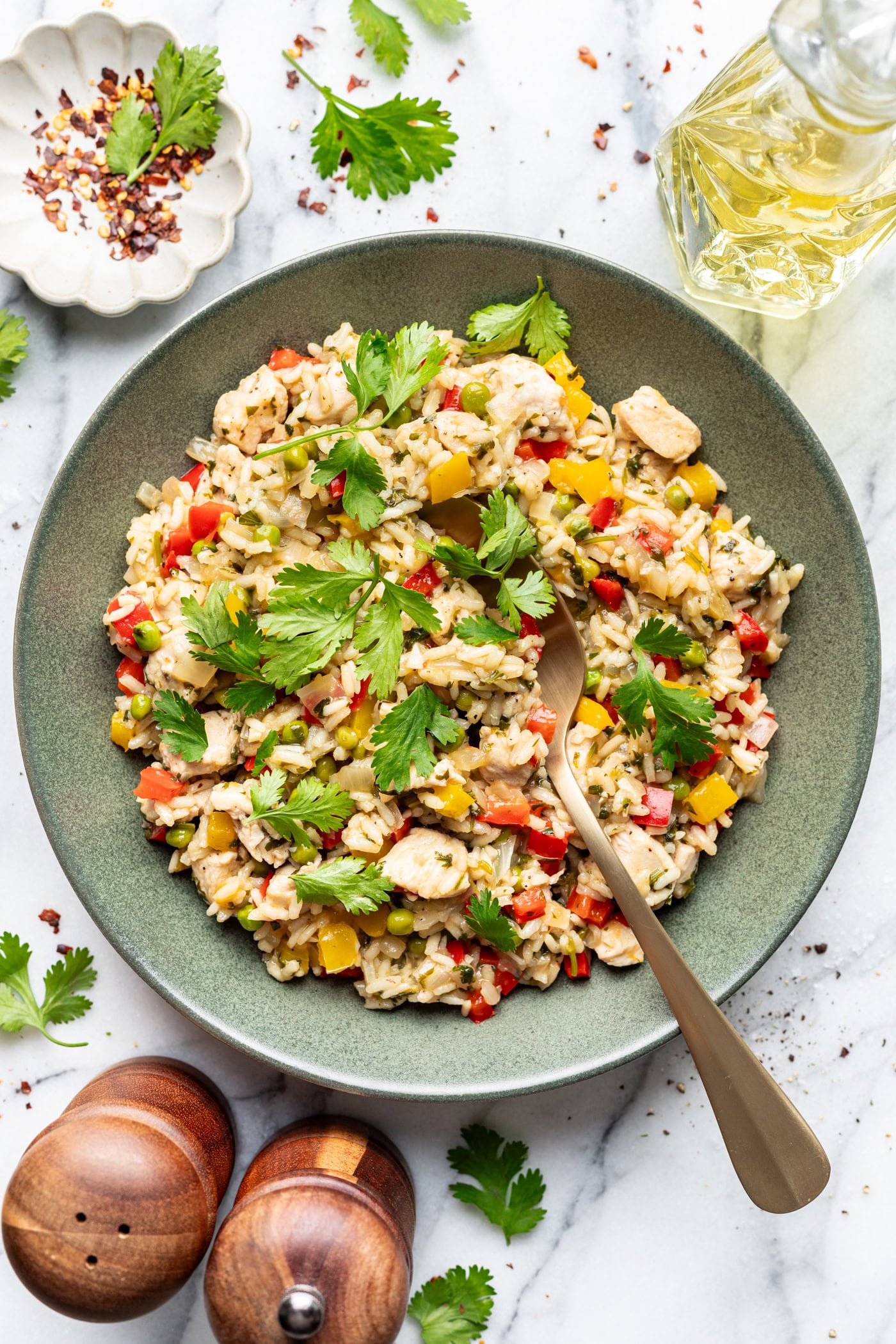 A green bowl filled with rice, chopped chicken, red and yellow bell peppers, and fresh cilantro, with a gold fork. Nearby are a small dish of red pepper flakes, a bottle of oil, cilantro leaves, and salt and pepper shakers.