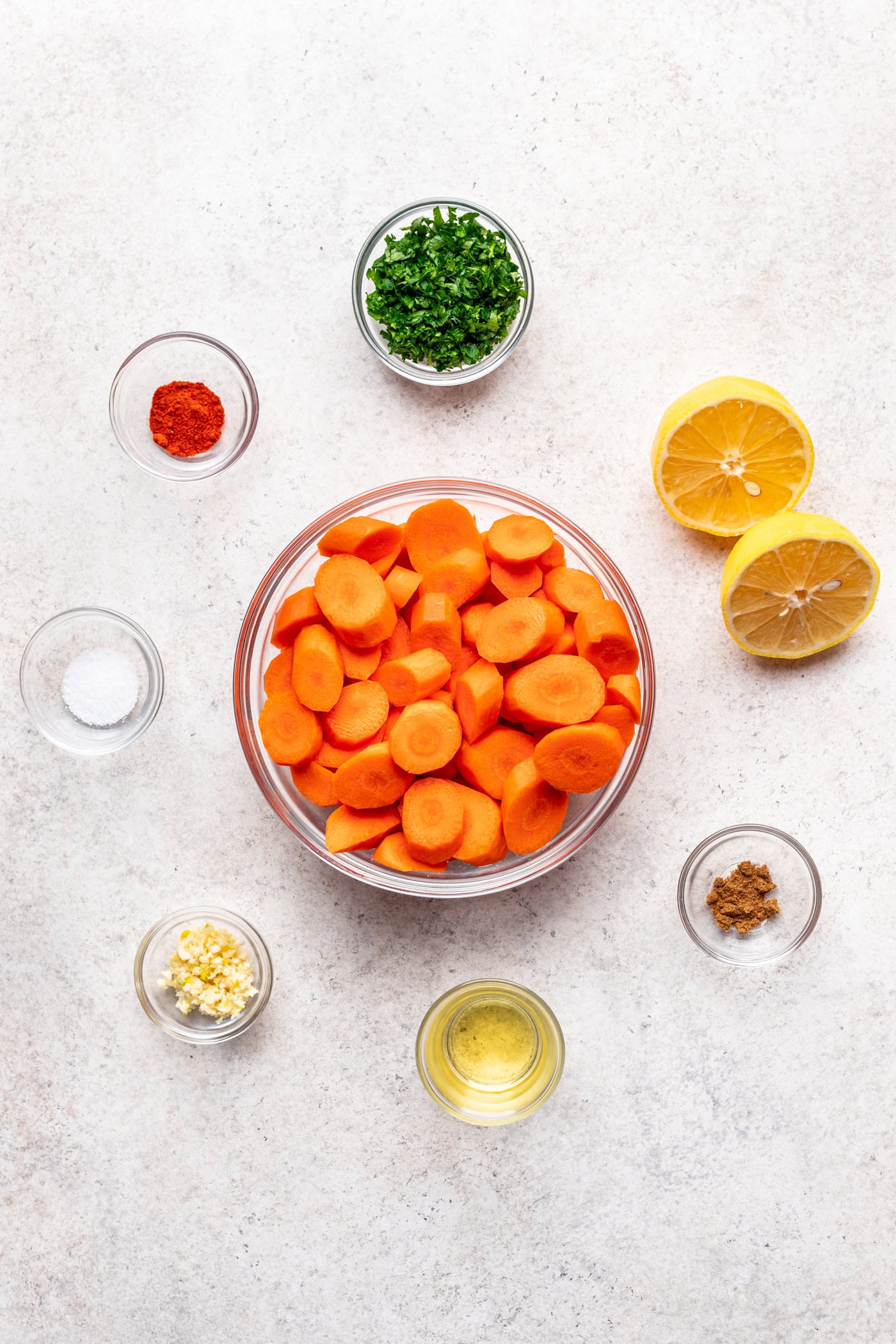A bowl of sliced carrots is surrounded by small bowls of minced garlic, chopped herbs, ground spice, paprika, salt, olive oil, and two halves of a lemon, all arranged on a light surface.