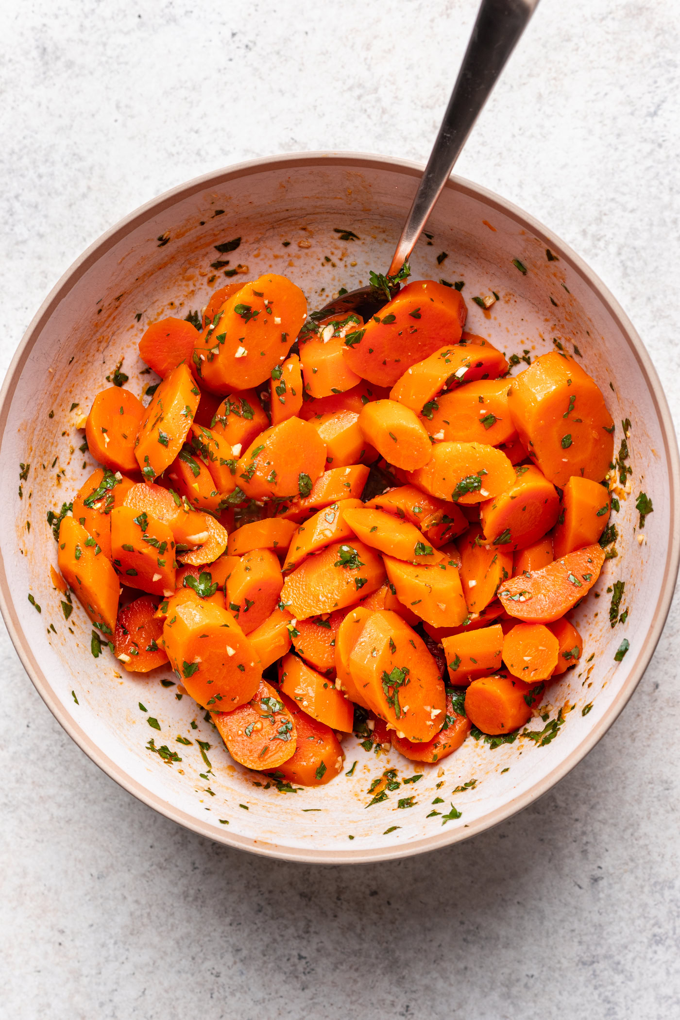 A bowl of sliced carrots mixed with chopped herbs and seasoning, with a spoon resting inside the bowl, set on a light background.