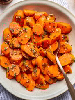 A bowl of sliced cooked carrots garnished with chopped herbs and minced garlic, served with a spoon on a light, striped cloth.