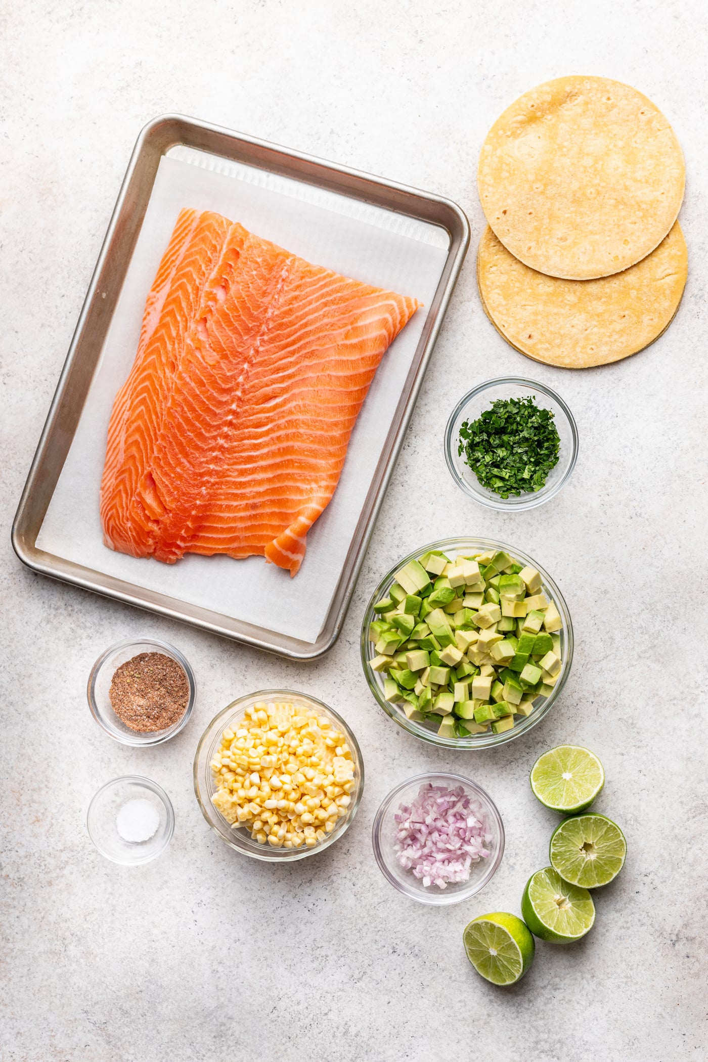 Overhead view of recipe ingredients: a tray with raw salmon fillet, three corn tortillas, bowls of diced avocado, chopped cilantro, corn, diced shallot, seasoning, salt, and three halved limes on a light surface.