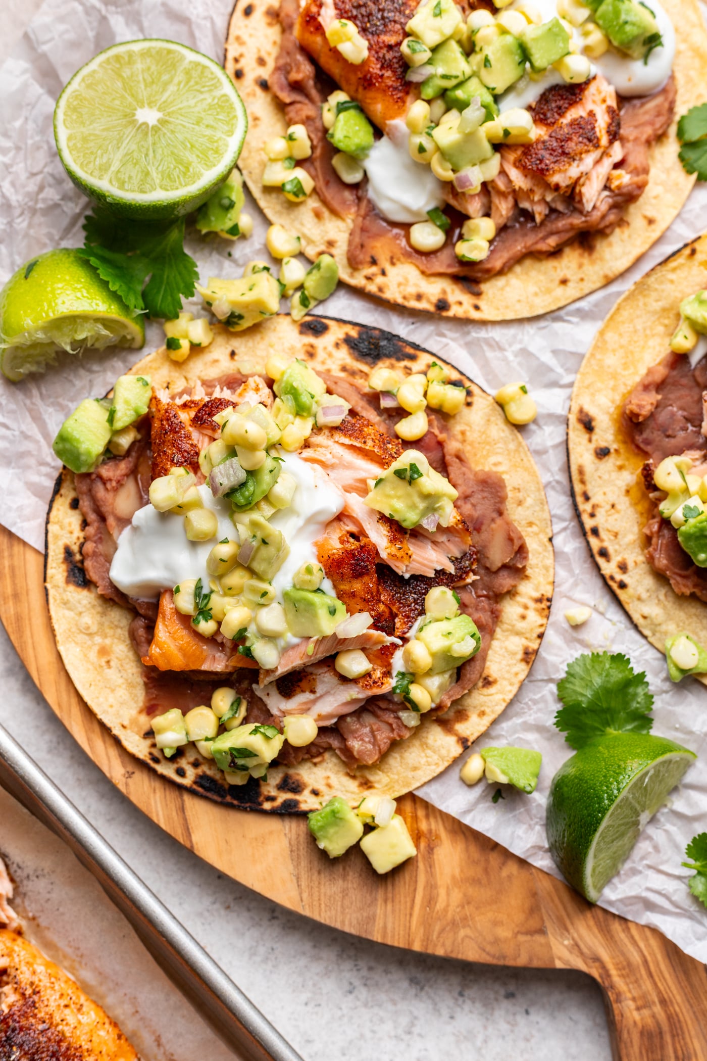 Three corn tortillas topped with refried beans, grilled salmon, white sauce, and a corn-avocado salsa, garnished with fresh cilantro and lime wedges on a wooden serving board.