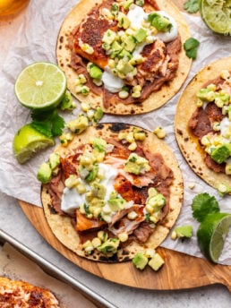 Three tostadas topped with refried beans, grilled fish, avocado salsa, and sour cream are garnished with cilantro and lime wedges on a wooden board covered with parchment paper.