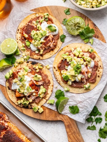 Three corn tostadas topped with refried beans, fish, corn salsa, avocado, and sour cream are arranged on a wooden board with lime wedges and cilantro. A bowl of salsa, fresh limes, and herbs are nearby.