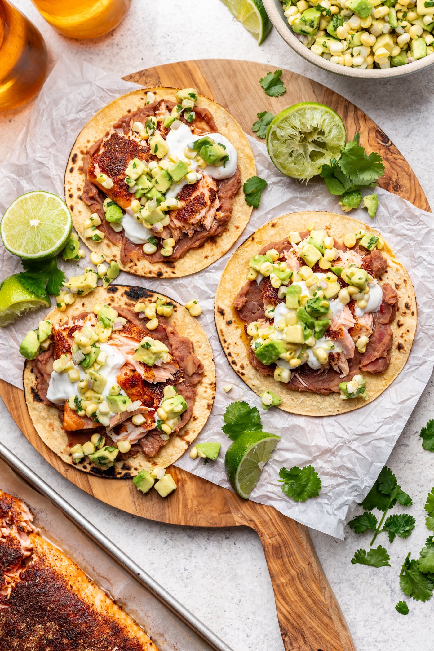 Three corn tostadas topped with refried beans, fish, corn salsa, avocado, and sour cream are arranged on a wooden board with lime wedges and cilantro. A bowl of salsa, fresh limes, and herbs are nearby.