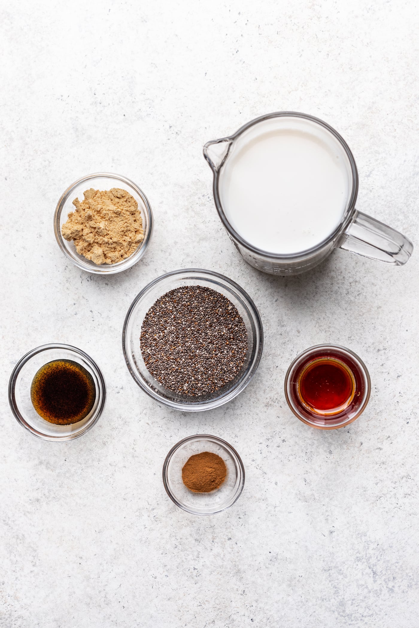A top-down view of six clear containers holding different ingredients: milk, chia seeds, peanut butter powder, vanilla bean paste, maple syrup, and ground cinnamon, arranged on a light textured surface.